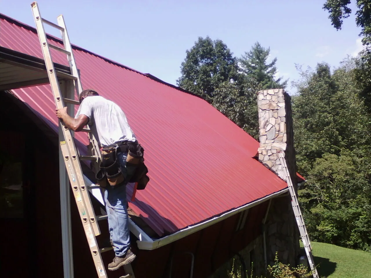 Roofer on a ladder beside a red metal roof and stone chimney — Soffit & Fascia Repair by Roofing Brothers in Payson